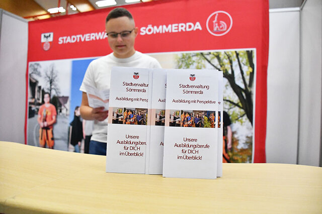 Vor dem Start der BIB 2025 am Stand der Stadt Sömmerda. Vor einem großen Plakat der Stadt Sömmerda steht ein Counter aus hellem Holz. Auf diesem sind zwei Flyer hochkant aufgestellt. In ihnen werden Ausbildungsberufe bei der Stadt Sömmerda vorgestellt. Hinter dem Counter steht ein junger Mann, im weißen T-Shirt und mit Brille ud blättert durch einen der Flyer.