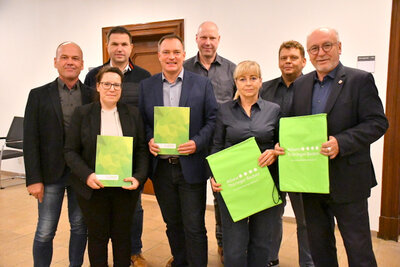 v. l.: Daniel Ecke (Weißensee), Julia Ansorg (Projektmanagerin), Hendrik Blose (Landgemeinde Buttstädt), Roman Zachar (Landgemeinde Kindelbrück), Dirk Barthels (Straußfurt), Beatrix Winter (Rastenberg), Uwe Kraneis (Kölleda), Ralf Hauboldt (Sömmerda Zwei Frauen und sechs Männer stehen in zwei Reihen hintereinander im Foyer des Rathauses. Die zwei frauen und zwei Männer in der ersten Reihe halten große Umschläge bzw. Rucksäcke im Hellgrün der Allianz in den Händen, auf denen das Logo der Allianz - vier Punkte und dazu der Schriftzug Allianz Thüringer Becken - steht.