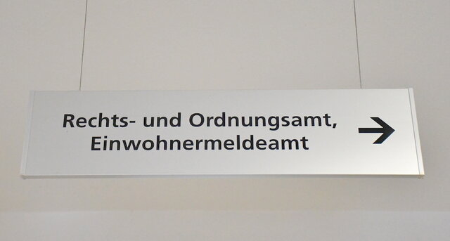 Das Schild im Verwaltungsgebäude Poststraße, welches auf das Einwohnermeldeamt verweist. Von der Decke hängendes, rechteckiges Hinweisschild aus gebürstetem Edelstahl im Verwaltungsgebäude Poststraße. In der oberen Zeile steht Rechts- und Ordnungsamt, darunter das Wort "Einwohnermeldeamt".