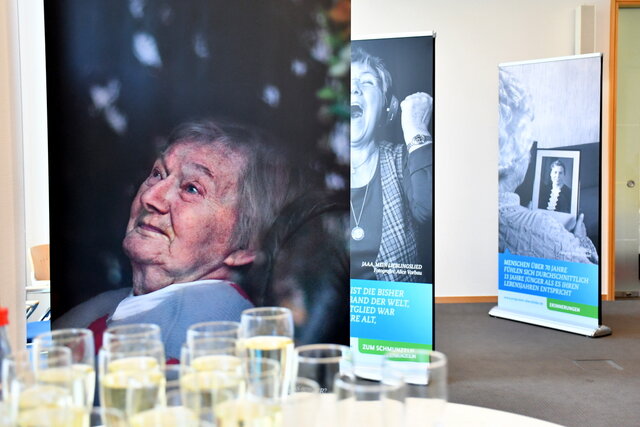 Ältere Frau als Porträt auf einem Roll-Up, dahinter stehen noch zwei Plakate die zur Ausstellung gehören. Auf beiden Plakaten befinden sich ältere Menschen und weisen auf die herrschenden Altersbilder hin