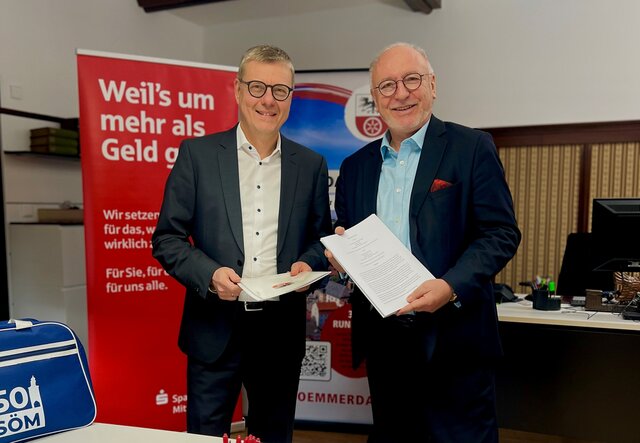 Unterzeichnung des Sponsoringvertrags mit der Sparkasse Mittelthüringen Bürgermeister Ralf Hauboldt und Sparkassen-Vorstandsvorsitzender Hans-Georg Dorst stehen im Rathaus nebeneinander und halten den frisch unterzeichneten Sponsoringvertrag für das Stadtjubiläum 2026 in den Händen. Im Hintergrund sind Werbebanner der Sparkasse und der Stadt zu sehen.