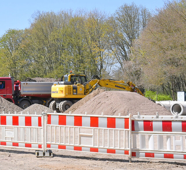 Rot-weiße Absperrbaken stehen vor zwei Baufahrzeugen und einem Hügel aus Sand bzw. Kies.