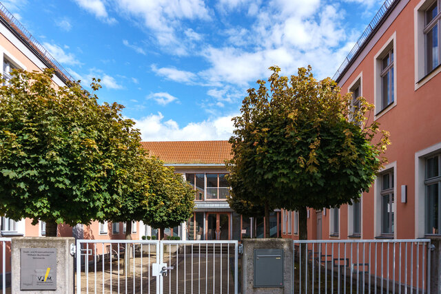 Blick in den Innenhof der Musikschule mit dem Haupteingang. Die Musikschule in SÖmmerda von außen. Es ist ein u-förmiger Gebäudekomplex in Dunkelrosa. Vom Eingang von der Straßenseite her blickt direkt auf den Haupteingang der Musikschule. Im Innenhof der Musikschule stehen links und rechts des Hauptweges Bäume.