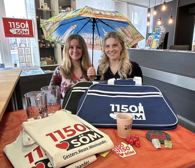 Julia Eberhardt (l.) und Nora Kästner präsentieren die Souvenirs. Ein kleiner Tisch in der Tourist-Information ist bedeckt mit Souveniers: Taschen, Beutel, Würfelbecher mit Würfeln, Pins, Stocknägel Untersetzer und Schirmen. Dahiner die Gesicher der zwei Mitarbeiterinnen, die einen Schirm mit Sömmerda-Motiven über sich aufgespannt haben.