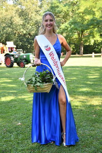 Celina steht im Stadtpark in einem blauen Kleid mit einem Waidenkorb in der Hand mit einer Shärpe und dem Aufdruck Sömmerdaer Waidprinzessin