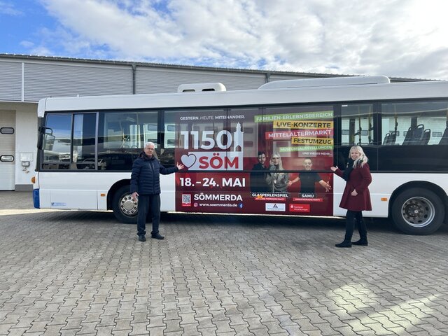 Der Linienbus mit der Werbung fürs Stadtjubiläum und die Festwoche. Auf diese weisen Olaf Silge, Geschäftsführer der Verwaltungsgesellschaft des ÖPNV, sowie Laura Scherzberg, Abteilungsleiter Kultur und Tourismus bei der Stadtverwaltung, hin. Ein weißer Bus ist von außen in der Mitte mit Werbung - ähnlich einem Plakat - zum Stadtjubiläum gestaltet. Vor dem Bus stehen links vor dem Plakat Olaf Silge, Schäftsführer der Verwaltungsgesellschaft des ÖPNV, und rechts Laura Scherzberg, Abteilungsleiterin Kultur und Tourismus bei der Stadtverwaltung. Beide zeigen auf die großflächige Werbung auf dem Bus.
