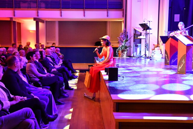 Impression vom Neujahrskonzert 2025 - damals mit dem Orchester Franz 'L. Eine Frau mit rotem Kleid und Basecap sitzt am Rand der Bühne im Volkshaus und singt dem Publikum im Saal zugewandt, das von seinen Stuhlreihen aus auf die Frau und die Bühne blickt. Auf der Bühne hat die Kamera zwei Notenpulte, an denen Franz 'L geschrieben steht. Vom kleinen Orchester wird im Bildausschnitt eine Geigen-Spielerin im roten Kleid gezeigt, die gerade auf ihrem Instrument spielt.