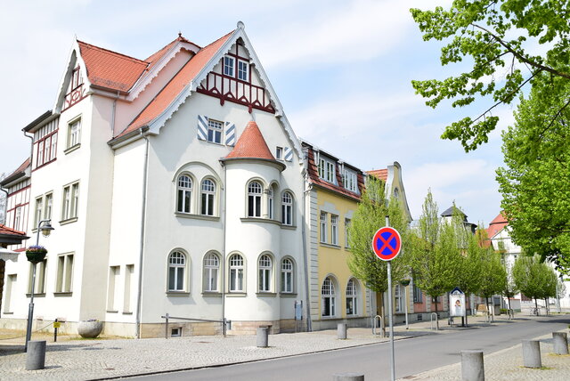 Volkshaus Sömmerda – Außenansicht Straßenansicht des Volkshauses Sömmerda in der Weißenseer Straße. Das helle Gebäude mit roten Dachziegeln und verzierten Giebeln liegt an einer gepflasterten Straße, die von Bäumen gesäumt ist. Im Vordergrund steht ein Verkehrsschild mit Halteverbot.