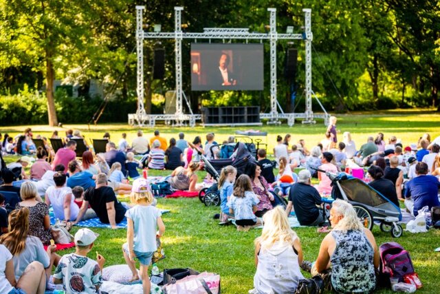 Titel: KJP-Familiensommerkino im Stadtpark Sömmerda Viele Besucherinnen und Besucher sitzen bei sommerlichem Wetter auf Decken und Campingstühlen im Stadtpark Sömmerda. Im Hintergrund ist die Leinwand des KJP-Familiensommerkinos zu sehen.