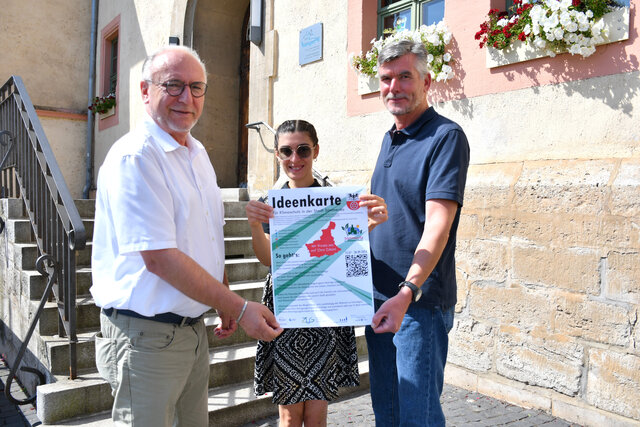 Bürgermeister, Klimaschutzmanagerin und der Leiter des Bau- und Umweltamtes zeigen vor dem Rathaus das Plakt zur Bürgerbeteiligung.