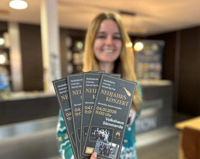 Auf den Flyern, die in der Tourist-Information ausliegen, gibt es die wichtigsten Informationen zum Neujahrskonzert. Eíne junge Frau hält Flyer in die Höhe. Auf diesen gibt es Hinweise zum Neujahrskonzert sowie zwei Fotos vom Mercurius-Sextett Dresden. Hinter der jungen Frau blickt man auf den Verkaufstresen und Auslagen der Tourist-Information.