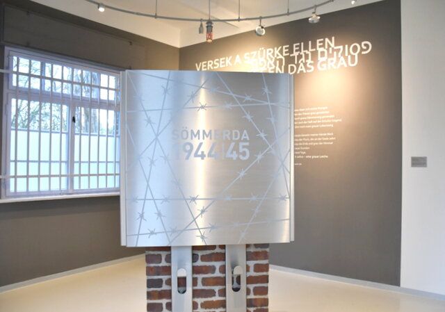 Blick ins Innere des Gedenk- und Bildungsortes "Tor 8" mit dem Mahnmal - einem Kubus aus Edelstahl. Im Inneren des gedenk- und Bildungsortes "Tor 8" steht auf einem Backsteinsockel ein Kubus aus Edelstahl. Die Vorderseite zeigt eingelasertes Stacheldraht als Zeichen für den ehemaligen Außenstandort des KZ Buchenwald in Sömmerda. In der Mitte der Vorderseite stehen das Wort Sömmerda und die Jahreszahlen 1944 und 45. Im Hintergrund steht an der grauen Wand mit weißen Buchstaben das Gedicht "Grau". Verfasst hat es während ihrer Zeit im Außenstandort Sömmerda des KZ Buchenwald eine der jüdischen Häftlingsfrauen. Die Überschrift des Gedichtes ist in Ungarisch, Hebräisch und Deutsch angebracht.