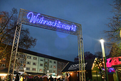 Blick auf den Eingangsberiech des Weihnachtsmarktes in den Abendstunden. Im Eingangbereich des Weihnachtsmarktes leuchtet der Schriftzug Weihnachtsmarkt in blauer Farbe. Dahinter wird der Blick frei auf verschiedene Hütten und Besucher des Weihnachtsmarktes sowie Häuser, die den Obermarkt begrenzen, freigegeben.