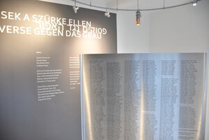 Einblick in den Innenraum des Mahnmals mit der Gedenktafel Metallische Gedenktafel mit den eingelserten Namen aller Inhaftierten welche vor einer braun farbenen Wand mit einem Gedicht steht.