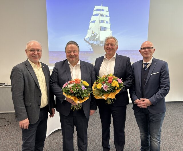 v. l.: Ralf Hauboldt, Klaus-Dietrich Matuschek, Uwe Kreitel und Andreas Böffel. Vier Männer stehen in einer Reihe nebeneinander. Dabei handelt es sich um Ralf Hauboldt, Klaus-Dietrich Matuschek, Uwe Kreitewl und Andreas Böffel. Herr Matuschek und Herr Kreitel halten je einen Blumenstrauß in der Hand. Im Hintergrund ist mit einem Beamer ein großes Segelschiff an die Wand projeziert. Es soll sinnbildlich für das Schiff SEV stehen.