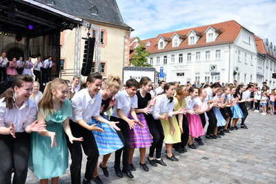 Der Tanzsportverein bei einem seiner Auftritte zum Stadtfest. Jugendliche Tänzerinnen und Tänzern tanzen in einer Reihe vor der Bühne. Auf der Bühne tanzen weitere Mitglieder der Gruppe. Den Platz säumt Publikum, welches bei der Aufführung zuschaut.
