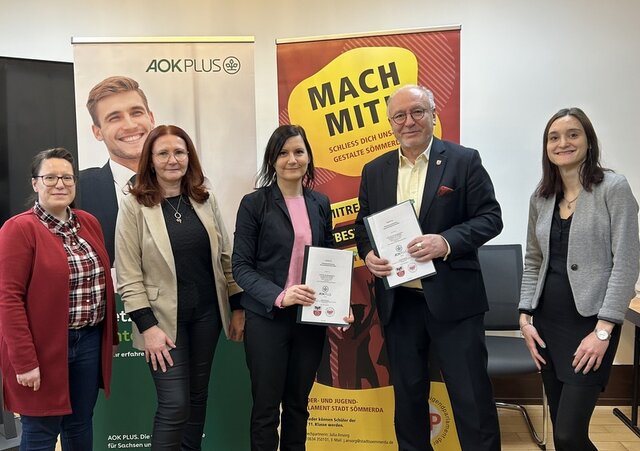 v. l.: Julia Ansorg, Ansprechpartnerin KJP bei der Stadtverwaltung, Chris Roßmeyer (Key Account Mangerin AOK plus), Nicole Koch (Leiterin Regionalcenter AOK plus), Bürgermeister Ralf Hauboldt sowie Susann Trautmann (Leiterin Kundencenter-Standort Sömme Drei Frauen von der AOK PLUS, die zuständige Mitarbeiterin der Stadtverwaltung und der Bürgermeister stehen in einer Reihe. Bürgermeister und eine Frau von der AOK PLUS halten jeweils ein Exemplar des unterzeichneten Vertrags zur Verlängerung der Gesundheitspartnerschaft in der Hand. Im Hintergrund stehen ein Rollup des Kinder- und Jugendparlaments der Stadt Sömmerda sowie der AOK PLUS.