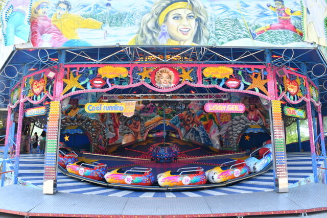 Am Stadtpark ist wieder Frühlingsfest. Ein größeres Fahrgeschäft auf dem Frühlingsfest. Es handelt sich um eine Art Karussell für Erwachsene. Links stehen außerhalb des Fahrgeschäftes einige Leute.