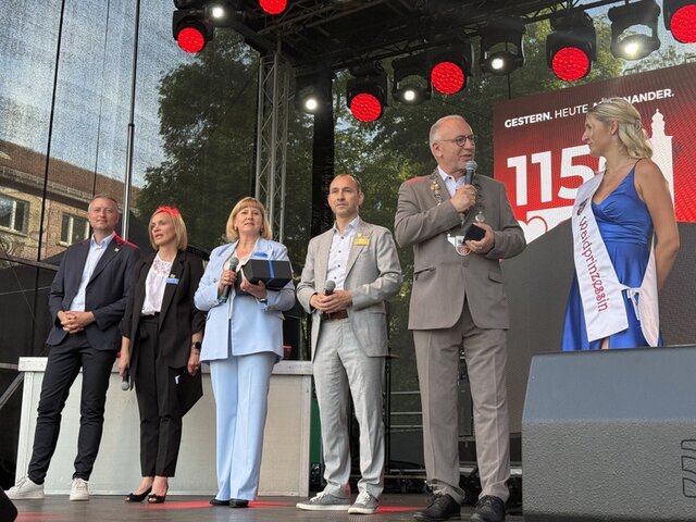 Auf der Bühne am Festwochenenende: Landrat, Dolmetscherin, stellvertretende Bürgermeisterin von Kedainiai, Oberbürgermeister von Böblingen, Sömmerdas Bürgermeister sowie die Waidprinzessin (v. l.). Auf der Bühne beim Stadtjubiläum stehen der Landrat sowie die stellvertretende Bürgermeisterin von Kedainiai, die Dolmetscherin und der Oberbürgermeister von Böblingen in einer Reihe. Leicht nach vorn gerückt steht Sömmerdas Bürgermeister mit der Sömmerdaer Waidprinzessin. Sie trägt ein langes blaues Kleid, eine weiße Schärpe mit roter Schrift sowie ein Diadem im blonden Haar. Bürgermeister und Waidprinzessin führen ein kurzes Gespräch über Mikrofon.