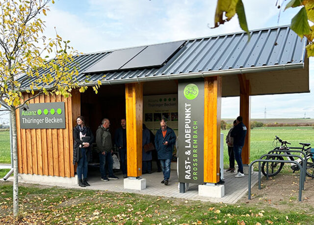 Der Rastplatz mit seiner Außenansicht. Ein überdachter Holzbau mit Solarpaneelen auf dem Dach. An dem Holzbau sind links und rechts Tafeln angebracht. Links mit dem Logo des Zweckverbandes Allianz "Thüringer Becken". Rechts mit dem Hinweis auf den rast- ud Ladepunkt Großbrembach. Zwei Fahrräder stehen am Holzbau. Unter dem Dach stehen mehrere Leute.