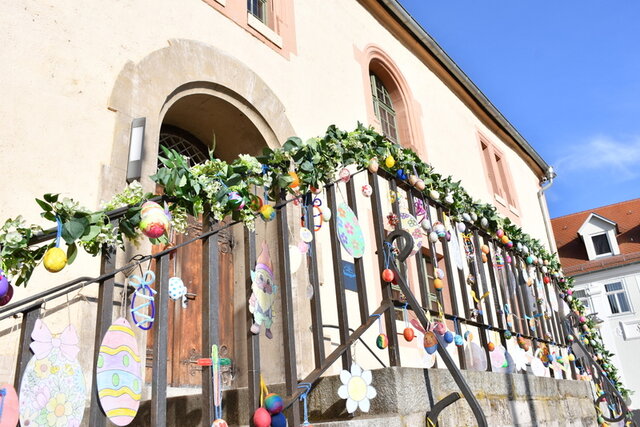 Geschmückter Aufgang zum Rathaus im Jahr 2025. Das Geländer am beidseitgen Treppenaufgang zum Rathaus-Eingang ist am Handlauf mit einer grünen Girlande umwunden. Darin hängen Ostereier in verschiedener Ausführung: bemalt oder aus starkem Papier und bunt bemalt. Die Sonne scheint und der Himmel leuchtet blau.