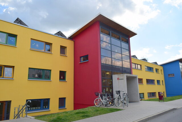 Auch die städtische Kindereinrichtung "Mischka" bleibt am 09. Januar geschlossen. Das Gebäude, das die städtische Kita "Mischka" sich mit der Evangelischen Grundschule teilt. Ein neuerer Bau mit vorgezogenem und an der Frontseite komplett verglastem Eingangsportal. Das dreistöckige Gebäude ist farblich in Gelb und Rot gehalten. Davor sind eine Zufahrt und eine schmale Rasenfläche zu sehen.