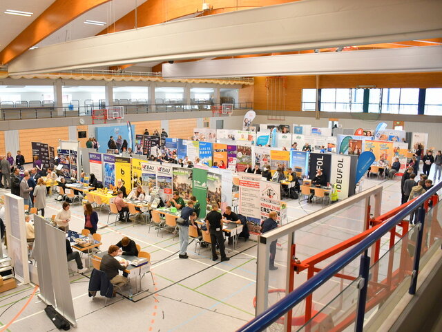 Draufsichts aufs Geschehen der Prakttikums- und Ausbildungsbörse 2025. In der großen und hellen Unstruthalle - eine Sport- und Mehrzweckhalle - stehen in mehreren Reihen Stände verschiedener Unternehmen und Institutionen nebeneinander. Hinter den Tischen mit den Werbebannern der Unternehmen sitzen Firmenvertreterinnen und Firmenvertreter. Vor den Tischen - allerdings nicht vor allen, sitzen junge Leute. Sie unterhalten sich mit den Firmenvertreterinnen und Firmenvertretern.