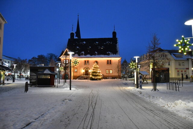 Das beleuchtete Rathaus mit Baum in der Adventszeit. Das mit Lichtkern erleuchtete Rathaus in der Dämmerstunde mit dem schöm beleuchtetwen Weihnachtsbaum davor. Wir blicken aus der Marktstraße aus einigem Abstand direkt auf Rathaus und Weihnachtsbaum. An den Straßenlampen hängen leuchtende Sterne. Es liegt eine leichte Schneedecke.
