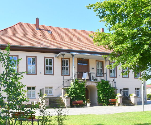 Das sanierte Gutshaus in Tunzenhaus. Über eine Wiese hinweg und zwischen zwei Bäumen hindurch geht der Blick auf das sanierte Gutshaus in Tunzenhausen. Es ist ein sonniger Tag mit blauem Himmel. Das Gutshaus ist ein zweistöckiges helles Gebäude. Zur Eingangstür führt ein von zwei Seiten begehbarer Treppenaufgang. Vor dem Treppenaufgang steht, getrennt durch einen Gewölbeeingang im Erdegeschoss, jeweils eine Sitzbank. Dahinter ranken sich Kletterpflanzen bis zum Geländer des Treppenaufgangs-Plateau hinauf.