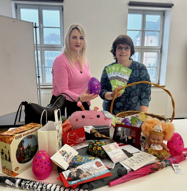 Lena Kob (l.) und Katrin Ewald bei der Ziehung der Preisträger mit einem der versteckten Ostereier, den Flyern zum Gewinnspiel sowie gestifteten Preisen. Zwei Frauen stehen hinter einem Tisch. Eine der Frauen hält präsentier ein größeres farbiges Osterei aus Plastik. Es ist eines von den Eiern, die in den Geschäften versteckt wurden. Die andere Frau hält einige der Gewinnkarten, die gezogen wurden, in der Hand. Auf dem Tisch vor ihnen liegen bzw. stehen die Gutscheine und Sachpreise, die die Geschäfte gestifet haben.