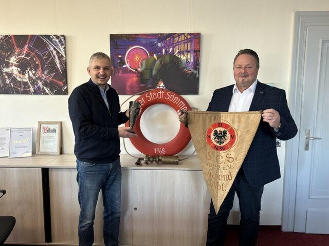 Dr. Hans-Diether Dörfler (l.) übernimmt die vier Objekte aus den Händen von Klaus-Dietrich Matuschek. Herr Dörfler und Herr Matuschek stehen in einem Raum vor einem flachen Schrank. Herr Dörfler hält einen Pokal in den Händen, Herr Matuschek einen Wimpel. An der Wand lehnt der rote Rettungsring. Hinter den beiden Männern hängen Bilder an der Wand.