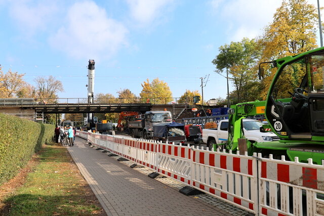 Blick auf den Baustellenbereich von der Innenstadt kommend Die jetzige Baustellensituation von der Stadtmitte kommend: Der Fußweg im Baustellenbreich ist mit rot-weißen Baaken von der Straße abgetrennt. Hinter den Baaken stehen verschiedene Baufahrzeuge auf der Straße. Links neben dem Fußweg befindet sich eine Grünfläche. Eine kleine Gruppe Fußgänger steht in einer Entfernung der Brücke und schaut den Arbeitenden zu. Hinter der Brücke ragt ein großer Kran auf.