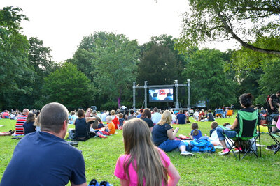 Familiensommerkino im Stadtpark. Viele Kinder, Jugendliche und Erwachsene sitzen in Gruppen verteilt auf Decken oder Campingstühlen auf einer großen Rasenfläche im Stadtpark, umgeben von Bäumen. Auf einer großen Leinwand wird ein Film gezeigt.