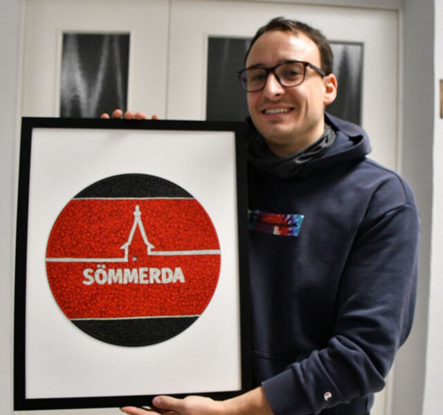 Erik Konrad Kästner, wisschenschaftlicher Mitarbeiter im Museum, mit einer der für die Mitmach-Ausstellung eingereichten Bildern. Ein junger Mann hält ein großes Bild hoch. In schwarzem Rahmen auf weißem Hintergrund zeigt es eine runde Scheibe in den farben schwarz und rot mit stilisierter Silhouette von Kirchturm und Stadtmauer. Der junge Mann trägt eine Brille und hat einen dunkelgrauen Kapuzenpullover an.