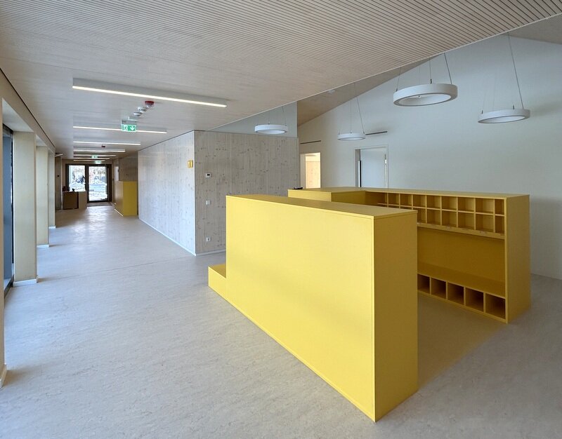 Blick in einen der weiträumigen Flure der Kita mit Garderobenbereich. Ein großer und breiter Flur der Kita. Links begrenzt durch bodentiefe Fenster. Rechts ist der Flur verkleidet mit hellem Holz. Im Vordergund steht eine Holz-Regalkombination in U-Form als Garderobe für die Sachen der Kinder. Die Garderobe ist in einem knalligen Gelb gehalten. Der Fußboden ist im Garderobenteil mit gelben Linoleum belegt. Der Rest des Flures in einem hellen Beigeton.