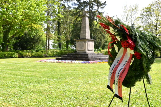Das Ehrenmal auf dem Friedhof im Hintergrund und der Kranz, der im vergangenen Jahr niedergelegt wurde. Eine grüne Rasenfläche mit einem großen steinernen Obelisken auf einer Treppenanlage drumherum. Um die Treffenanlage sind Blumen gepflanzt. Im Vordergrund rechts steht ein Gestell, dass den Kranz des Bürgermeisters hält. Der grüne Kranz ist bestück mit roten und weißen Kunstblumen sowie zwei rot-weißen Bändern. Auf ihnen steht folgendes: In ehrendem Gedenken Bürgermeister Stadt Sömmerda.