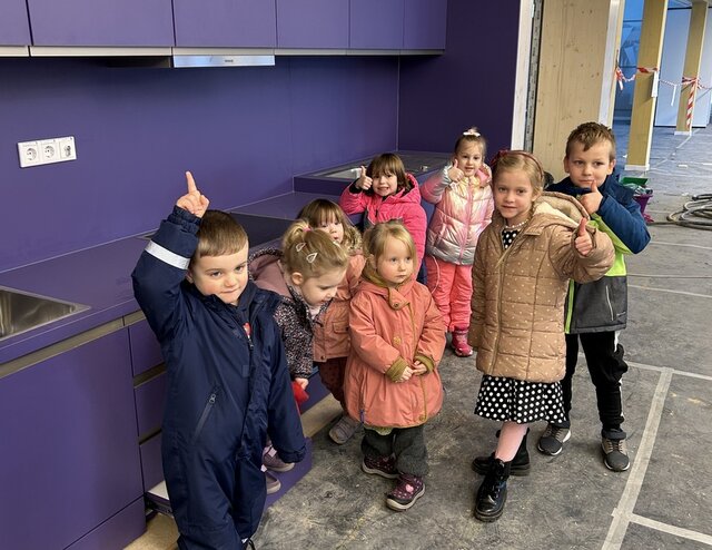 Kita-Kinder vor der Kinderküche im Neubau. Eine Kindergruppe steht im Neu ihrer Kita vor einer dunkellilafarbenen Kinderküche und winkt in Richtung Fotografin. Im Hintergrund des Fotos sind in regelmäßigen Abständen helle Holzpfeiler zu sehen. Zwischen ihnen wurde rot-weißes Band zur Absperrung gezogen. Der Fußboden ist zum Schutz vor Schmutz noch mit dünnem Filz ausgelegt.