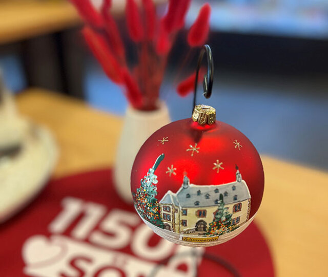 Die exklusive Weihnachtskugel mit Rathaus-Motiv und Weihnachtsbaum. Eine rote, matt glänzende Kugel hängt an an einem kleinen Ständer. Kleine goldene Sterne rahmen das auf die Kugel kunstvoll gemalte Sömmerdaer Rathaus und eines geschmückten Weihnachtsbaums. Im Hintergrund steht ein Strauß mit rot gefärbten Gräsern.