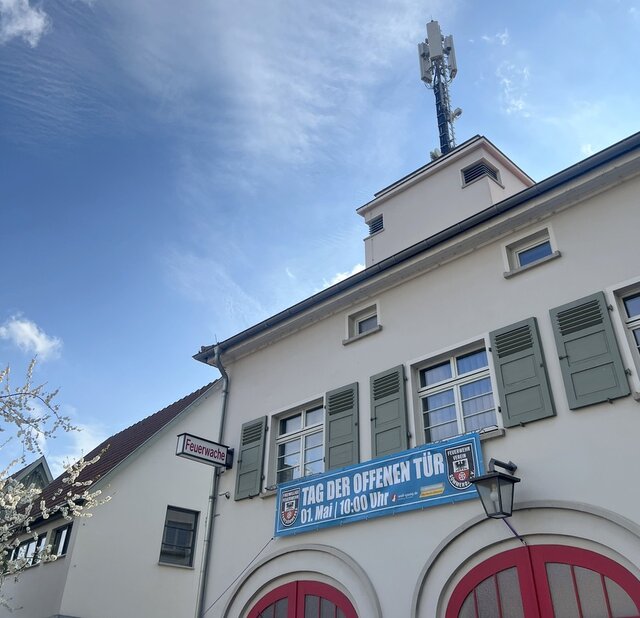 Das Banner an der Feuerwache weist auf den Tag der offenen Tür hin. Ein Teil eines Gebäudes der Feuerwache Sömmerda mit roten Rundbogen-Garagentüren sowie Sirenenanlage auf dem Dach. Und der oberen Fensterrreihe hängt ein Banner, das auf den Tag der offen Tür am 1. Mai hinweist.