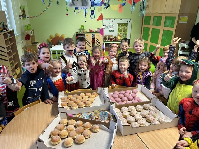 Auch in der Kita "Bummi" in Leubingen freuten sich Kinder über die Pfannkuchen. Auf einem Tisch in einem Kita-Gruppenraum stehen vier flache Kartons mit Pfannkuchen. Diese sind mit Zucker umhüllt oder haben eine weiße Glasur mit bunten Streuseln oder eine rosa Glasur. Um den Tisch herum stehen Kinder in Faschingskostümen, strecken den Daumen in die Höhe in freuen sich über die Pfannkuchen.