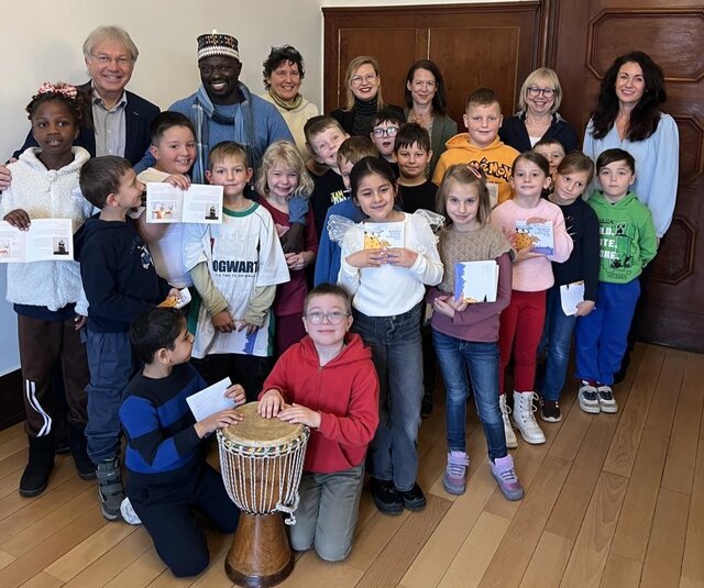 Eröffnung der 4. Thüringer Kinder- und Jugendliteraturtage mit der Klasse 2b im Rathaus Sömmerda Gruppe von Grundschulkindern der Klasse 2b der Diesterweg-Grundschule steht gemeinsam mit dem Geschichtenerzähler Ibrahima „Ibo“ Ndiaye, Mitarbeitenden der Stadt- und Kreisbibliothek sowie weiteren Erwachsenen für ein Gruppenfoto im Sitzungssaal des Rathauses Sömmerda. Einige Kinder halten Autogrammkarten und Programmhefte in der Hand, ein Kind sitzt vorne an einer Trommel. Alle lächeln in die Kamera.