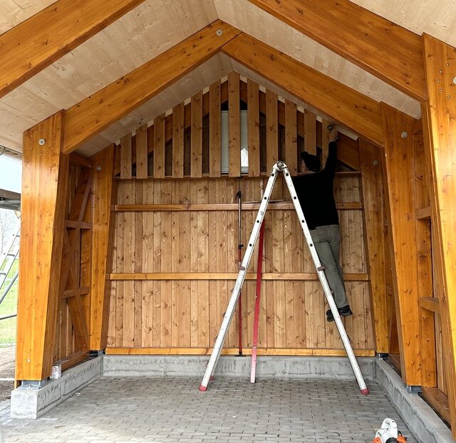 Der Radler-Rastplatz in Leubingen am Unstrut-Radweg ist noch in Bau. Ein offener Bau aus Holz. Ein Mann steht auf einer Leiter und schraubt bearbeitete Holzbretter an die Giebelseite des Carpot ähnlichen Baus.