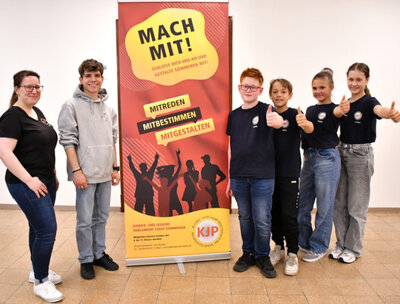 Werben für weitere Mitglieder im KJP: v. l. Julia Ansorg, Simeon, Louis, John, Meila und Hanna. Vier neue Mitglieder des Kinder- und Jugendparlaments stehen mit erhobenem Daumen rechts vom Plakat, auf dem für eine Mitgliedschaft im KJP geworben wird. Links vom Plakat stehen der KJP-Vorsitzende sowie die Ansprechpartnerin bei der Stadtverwaltung für das KJP.