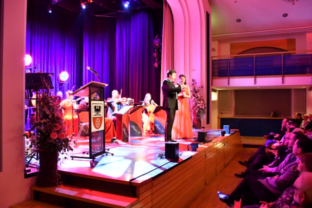 Im Volkshaussaal spielen die Musikerinnen und Musiker des Orchesters hinter schwarz-roten Notenpulten auf der Bühne auf ihren Instrumenten wie Geige, KOntrabass und Querflöte. Am Bühnenrand mit Blick in den Saal stehen die beiden Gesangssolisten und singen mit jeweils einem Mikrofon in der Hand. Das Fotos zeigt außerdem die erste Publikumsreihe im Volkshaussaal.