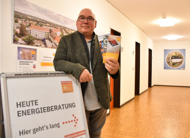 Frank Weber steht an einem Aufsteller in einem langen Flur und hält Flyer zur Energiebratung der Verbraucherzentrale in der linken Hand. Mit der rechten Hand stützt er sich auf den Aufsteller. An diesem ist ein Plakat angebracht, auf dem unter anderen steht: Heute Energieberatung - hier entlang. Auf dem Flur hängen an den Wänden Luftaufnahmen von Sömmerda.