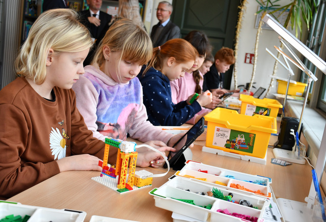 Kinder sitzen an einem langen Tisch. Sie stecken mit bunten Plastiksteinen aus einem Bausatzkasten verschiedene Objekte wie beispielsweise eine Schaukel zusammen und versuchen, mit Hilfe von Tablets die Objekte so zu programmieren, dass sie sich bewegen. Im Hintergrund stehen Erwachsene und unterhalten sich.