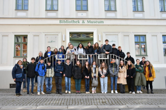 Die Schülerinnen und Schüler sowie die Lehrerinnen habe sich auf dem beidseitig zu begehenden Treppenaufgang zum Dreyse-Haus sowie vor dem Treppenaufgang zum Foto aufgestellt. Auf der hellen Fassade des sanierten historischen Dreyse-Hauses stehen in grüner Schrift die Worte Bibliothek & Museum.