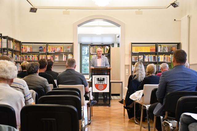 Ein Saal bestückt mit mehreren Stuhlreihen recht und links vom Mittelgang. Vorn am Rednerpunlt steht der Bürgermeister und hält seine Rede mit Blick ins Publikum.