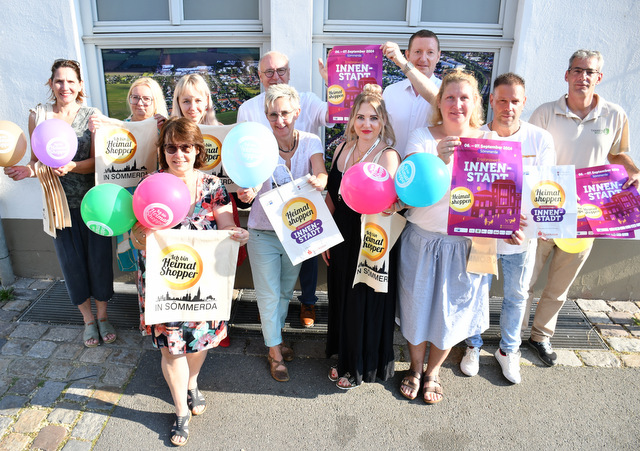 Eine Gruppe von Männern und Frauen steht vor einem Geschäft und hält Plakate, Luftballons und Gewinnspielkarten als Werbematerial fürs Heimat shoppen in die Höhe.