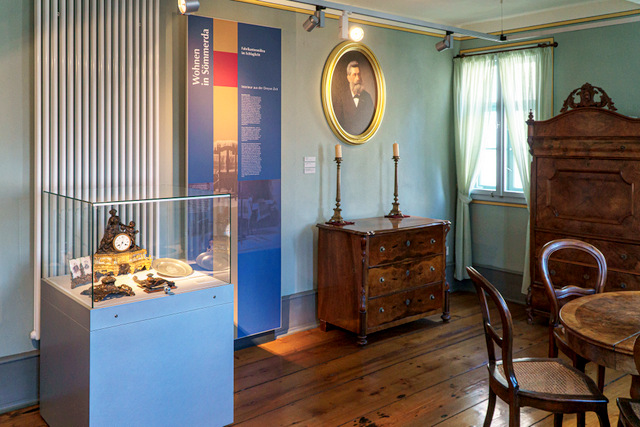 Blick in einen der Räume der historischen Ausstellung des Museums. Einer der Räume der historischen Ausstellung im Museum. In dem Raum stehen eine Vitrine mit kleineren Exponaten, eine Kommode, Schrank, Tisch und Stühle. An der Wand hängen eine Erläuterungstafel sowie das Porträt eines Mannes mit Bart.